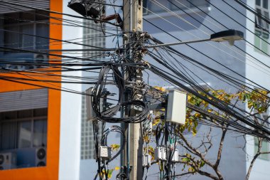Bir elektrik direğindeki karmaşık elektrik kablolarının ve kabloların yakın plan görüntüsü, şehir ortamındaki renkli modern binalarla tezat oluşturuyor..