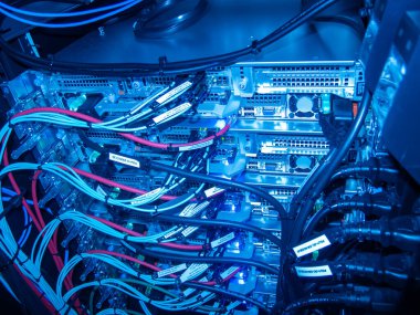 Bu resim, karmaşık kablolar ve devre kartlarıyla dolu bir sunucu rafının mavi tonlu görüntüsünü sergiliyor. Belgede veri yönetimi için gerekli teknoloji altyapısı vurgulanıyor.