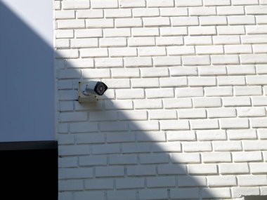 Beyaz tuğladan bir duvara monte edilmiş bir güvenlik kamerası güneş ışığına gölge düşürüyor. Bu görüntü modern mimaride kentsel güvenlik teknolojisi ve güvenlik tedbirlerini temsil etmektedir..