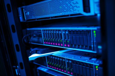 Mavi ışıklarla aydınlatılmış birden fazla veri depolama biriminin yer aldığı karanlık bir sunucu odasının yakın çekim görüntüsü. Teknoloji ve ağ ekipmanları modern dijital altyapıyı sergiliyor.