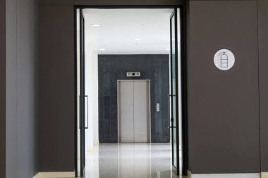 Bu resim, açık kapılar ve açık güvenlik levhalarıyla, minimalist ve profesyonel bir iç tasarımın ön plana çıkarıldığı modern bir asansör girişini gösteriyor..