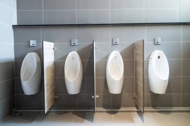 Dört beyaz pisuvarın bulunduğu modern bir tuvalet şık gri kiremitli bir duvara karşı. Bu tasarım kamu tesislerinde temizlik, hijyen ve işlevselliği vurgular..