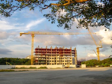 Yeni bir bina üzerinde çalışan bir vincin yer aldığı canlı bir inşaat alanı. Batan güneş, ağaçların çerçevelediği sahnede sıcak bir parıltı saçar..
