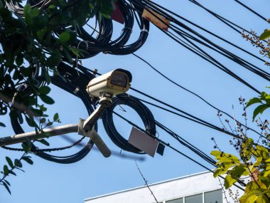 Bir güvenlik kamerası karmaşık elektrik kablolarının üzerinde, yeşilliklerle çevrili. Bu görüntü, kentsel alandaki teknoloji ve doğanın kesişimlerini yakalar..
