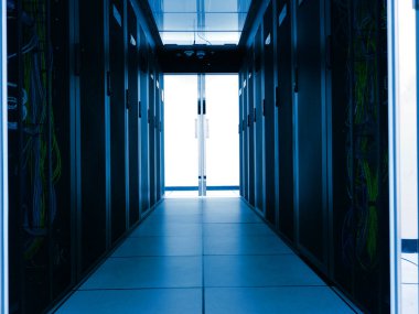 Sunucu odası içinde karanlık bir koridor, ağ dolapları ve renkli kablolar sergileniyor, parlak kapılara açılıyor. Teknoloji ve altyapı temaları için ideal.