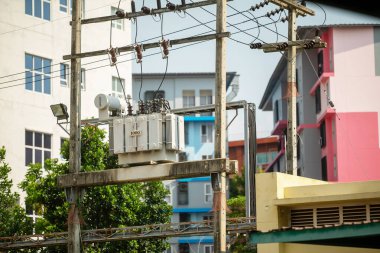 Şehir binaları arasında elektrik direğine monte edilmiş bir güç transformatörü. Görüntü, şehrin enerji dağılımı için gerekli olan elektrik altyapısını yakalar..