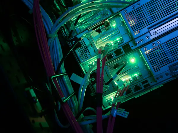 Ağ sunucu kablolarının ve aydınlatılmış teknoloji bileşenlerinin yakın görüntüsü. Parlak yeşil ve mavi ışıklar karmaşık bağlantıları ve elektronik altyapıyı vurgular.