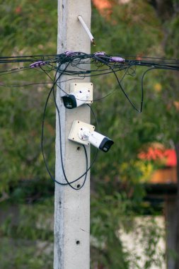İki güvenlik kamerası birbirine dolanmış kablolarla bir elektrik direğine monte edilmiş, güvenlik ve izleme sistemlerine odaklanmış kentsel bir çevre tasvir ediliyor.