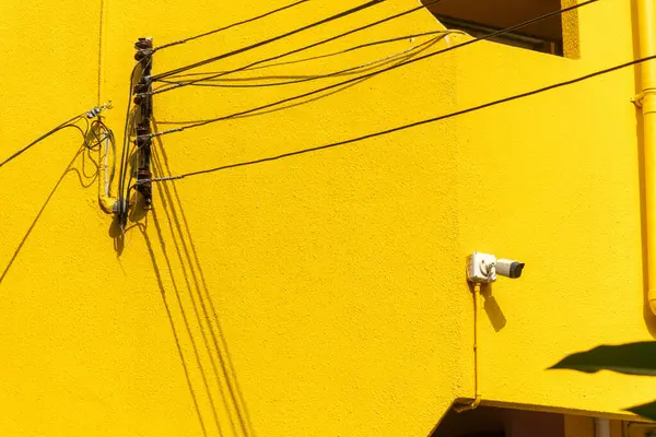 Bu görüntü, elektrik hatları ve güvenlik kamerasıyla süslenmiş canlı sarı bir duvar yakalıyor. Şehir tasarımı ve modern altyapı sergileniyor..
