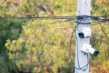 Elektrik kabloları ve güvenlik kameralarıyla dolu bir elektrik direğine yakın plan, yemyeşil bir zemin üzerine kurulmuş, kentsel altyapı sergileniyor..