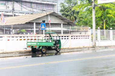 Yağmurlu bir caddeye park etmiş yeşil bir üç tekerlekli, şehir hayatını gözler önüne seriyor. Islak yol çevreyi yansıtıyor, canlı bir ortamda ulaşımı vurguluyor..