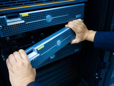 Bir teknisyenin elleri, modern bilişim altyapısı ve teknoloji bakımını vurgulayarak, veri merkezi rafında sunucu donanımı değiştirme sürecini göstermektedir..