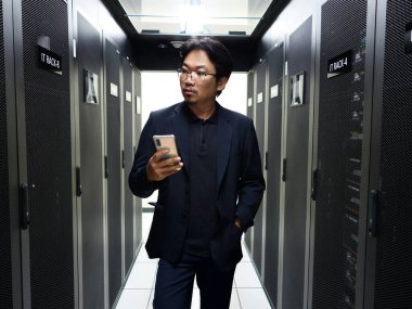 Bir iş adamı sunucu odasında mobil bir cihazla iletişim kurarak, kurumsal bir ortamda modern teknolojiyi ve dijital iletişimi sembolize eder..