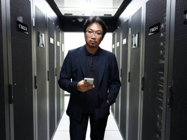 Resmi giyinmiş kendine güvenen bir adam veri merkezinde elinde akıllı bir telefonla dikiliyor. Görüntü, modern teknolojinin ve profesyonel iş ortamlarının özünü yakalıyor..