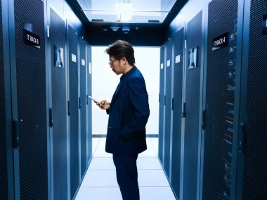 Takım elbiseli bir işadamı, sunucu raflarıyla çevrili modern bir veri merkezinin koridorunda dururken akıllı telefon etkileşimine girer..