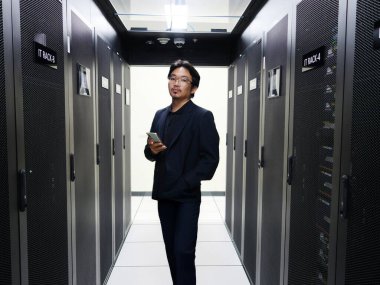 Profesyonel bir adam veri merkezinde elinde akıllı bir telefonla güvenle durur. Gösterişli ortamda modern bilişim çözümlerini vurgulayarak sunucu rafları ve ileri teknolojiler sergileniyor.