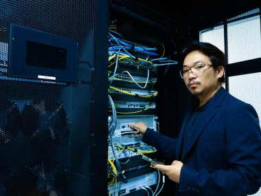 Bir teknisyen veri merkezinde ağ ekipmanlarını yönetiyor, kablo ve sunucuların düzgün çalışmasını sağlıyor. Sahne teknoloji profesyonelliğini vurguluyor.