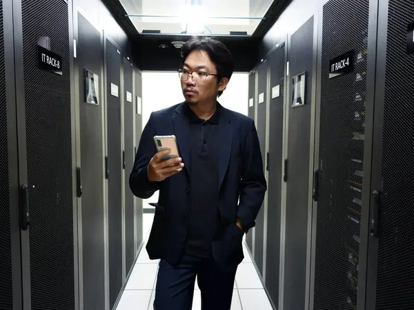 Bir iş adamı sunucu odasında mobil bir cihazla iletişim kurarak, kurumsal bir ortamda modern teknolojiyi ve dijital iletişimi sembolize eder..