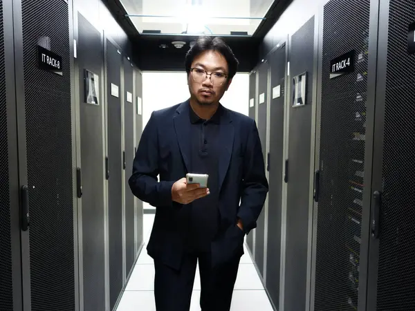 Resmi giyinmiş kendine güvenen bir adam veri merkezinde elinde akıllı bir telefonla dikiliyor. Görüntü, modern teknolojinin ve profesyonel iş ortamlarının özünü yakalıyor..