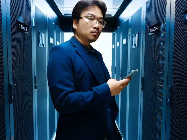 Profesyonel bir bilişim uzmanı bir sunucu odasında elinde akıllı bir telefonla güvenle durur. Atmosfer teknoloji ve veri yönetimini vurguluyor. İş ve teknoloji temaları için mükemmel.