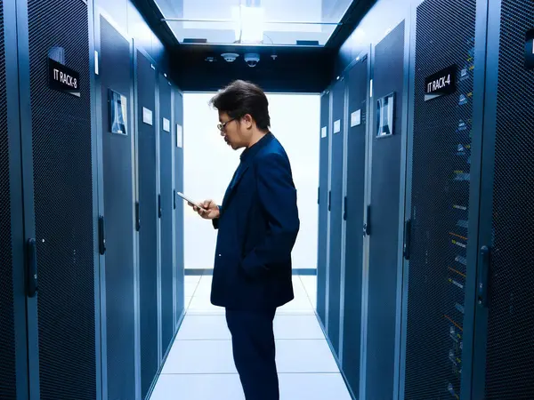 Takım elbiseli bir işadamı, sunucu raflarıyla çevrili modern bir veri merkezinin koridorunda dururken akıllı telefon etkileşimine girer..