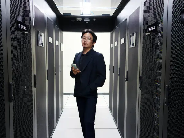 Profesyonel bir adam veri merkezinde elinde akıllı bir telefonla güvenle durur. Gösterişli ortamda modern bilişim çözümlerini vurgulayarak sunucu rafları ve ileri teknolojiler sergileniyor.