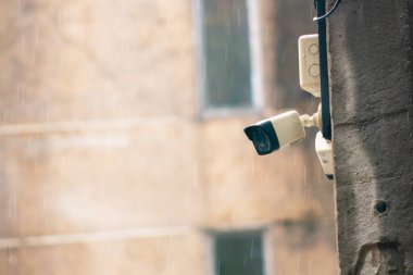 Duvara monte edilmiş bir güvenlik kamerası şehir güvenliğinin özünü yakalıyor. Kameranın etrafına yağan yağmur, teknolojinin ortamları izlemedeki rolünü vurguluyor.