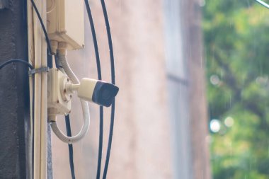 Duvara monte edilmiş bir güvenlik kamerası şehir ortamında düşen yağmuru kaydediyor. Bu görüntü, yıpranmış bir ortamda modern izleme teknolojisini vurguluyor..