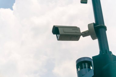 Görüntü, kabarık bulutlar ve mavi gökyüzü ile zıtlaşırken çevreyi yakalayan bir direğe monte edilmiş bir güvenlik kamerasını gösteriyor..