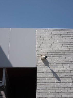 Beyaz tuğla duvara monte edilmiş bir güvenlik kamerası olan modern bir bina cephesi. Açık mavi gökyüzü mimari tasarıma derinlik katıyor..