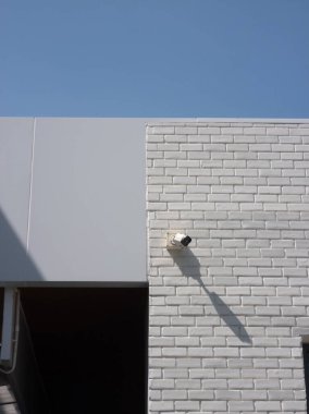 Beyaz tuğladan bir duvarın minimalist bir görüntüsü, üzerinde güvenlik kamerası olan, açık mavi gökyüzüne karşı, modern mimari ve tasarım unsurlarını vurgulayan,.