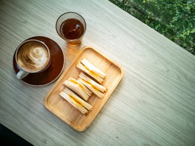 Rahatlatıcı bir içeceğin yanında nefis sandviçler ve bir fincan kahvenin bulunduğu ahşap bir tabakta rahat bir ortam, dinlenme anı için mükemmel..