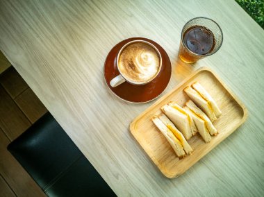 Bir fincan kahve, bir bardak soğuk içecek ve ahşap tepside bir tabak taze sandviç içeren samimi bir kahvaltı sahnesi..