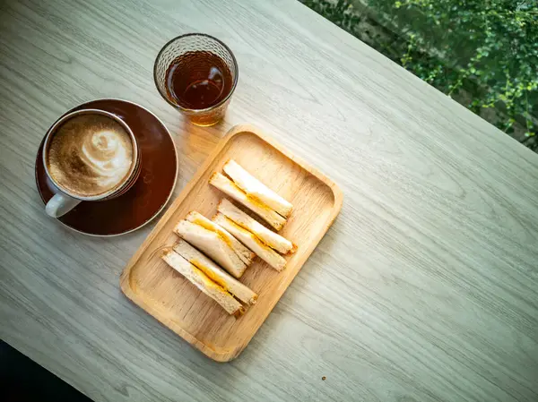 Rahatlatıcı bir içeceğin yanında nefis sandviçler ve bir fincan kahvenin bulunduğu ahşap bir tabakta rahat bir ortam, dinlenme anı için mükemmel..
