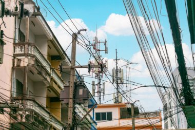 Binalar ve elektrik hatları arasında haberleşme kulelerinin yer aldığı canlı bir şehir manzarası, parlak mavi gökyüzü altında teknoloji ve şehir yaşamının kesişimini gösteriyor..