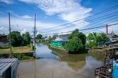 Berrak bir nehir manzarası yemyeşil ve mavi gökyüzünün yansımalarını yakalar. Elektrik hatları, sakin suların yanındaki evlerin görüntüsünü çerçeveleyerek manzaralı bir manzara yaratıyor..