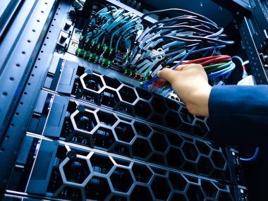 Bir sunucu rafındaki ağ kablolarını birbirine bağlayan bir elin yakınlaştırılması, bir bilişim ortamında modern teknoloji ve veri yönetiminin sergilenmesi.