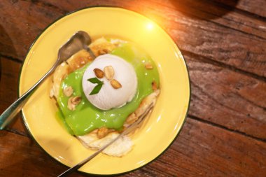 Yeşil sosla süslenmiş ve fındıkla süslenmiş yumuşak kreplerin tepesinde bir kepçe dondurmanın yer aldığı nefis bir tatlı..