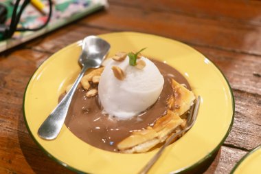 Zengin çikolata sosu ve fındıkla süslenmiş ve canlı sarı bir tabakta servis edilen bir kepçe dondurmanın yer aldığı enfes bir tatlı. Her durum için mükemmel.!