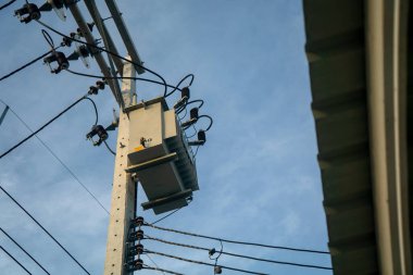 Görüntü, elektrik direğine monte edilmiş bir güç transformatörü gösteriyor. Elektrik kabloları temiz bir gökyüzüne dokunuyor ve modern elektrik altyapısını vurguluyor..