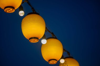 Koyu mavi bir gökyüzüne asılı sarı fenerlerin canlı bir görüntüsü, akşam toplantıları ve kutlamalar için sıcak ve davetkar bir atmosfer yaratıyor..