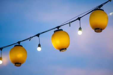 Parlak sarı fenerler zarif bir şekilde yumuşak mavi gökyüzüne asılıp açık hava toplantıları, festivaller ve kutlamalar için büyüleyici bir atmosfer yaratırlar..