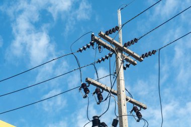 Bir elektrik direği, birden fazla kablosu ve bağlantıları olan parlak mavi bir gökyüzüne karşı dimdik durur. Bu görüntü modern enerji altyapısını ve güç dağılımını temsil ediyor..