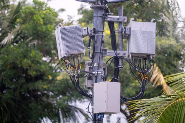 Telekomünikasyon sinyallerini yeşil bir ortamda arttırmak için tasarlanmış kablo ve antenleri gösteren elektrik direğine monte edilmiş iletişim ekipmanlarının yakın görüntüsü.
