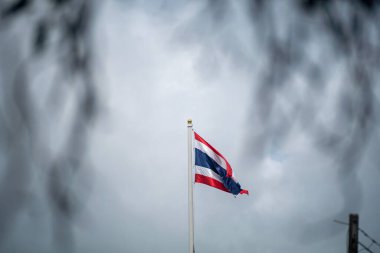 Canlı bir Tayland bayrağı bulutlu bir gökyüzüne gururla el sallıyor. Fotoğraf, ulusal kimliğin ve kültürel gururun özünü yansıtıyor..