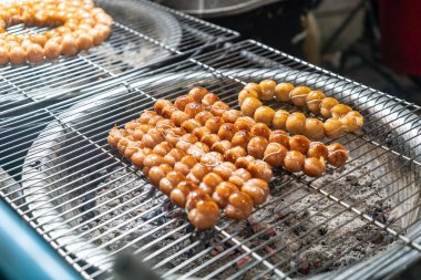 Mangal ızgarasında ızgara şiş ve sosis olan canlı bir gece pazarı sahnesi. Atmosfer, havada süzülen lezzetli aromalarla dolu..