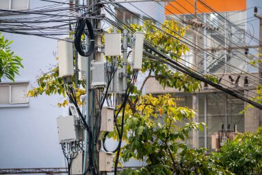 Çoklu elektrik kabloları ve kutuları olan bir elektrik direğini gösteren şehir manzarası. Görüntü, şehir manzarasının ortasındaki teknoloji ve doğanın kesişimini vurguluyor..