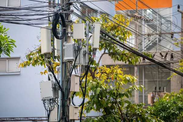 Çoklu elektrik kabloları ve kutuları olan bir elektrik direğini gösteren şehir manzarası. Görüntü, şehir manzarasının ortasındaki teknoloji ve doğanın kesişimini vurguluyor..