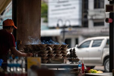 Bir pazar satıcısı yükselen dumanlar arasında sokak yemekleri hazırlıyor, yerel mutfağı ve canlı kültürünü sergiliyor. Sahne, şehir yaşamının ve gıda hazırlığının enerjisini yakalıyor..