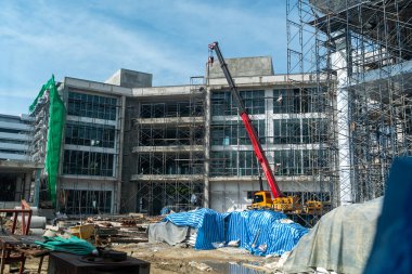 Bir inşaat sahasında, iskeleler ve koruyucu brandalarla çevrili vinç kaldırma malzemeleri sergileniyor. İşçiler, açık mavi gökyüzü altında inşaat geliştirme çalışmalarına giriştiler.